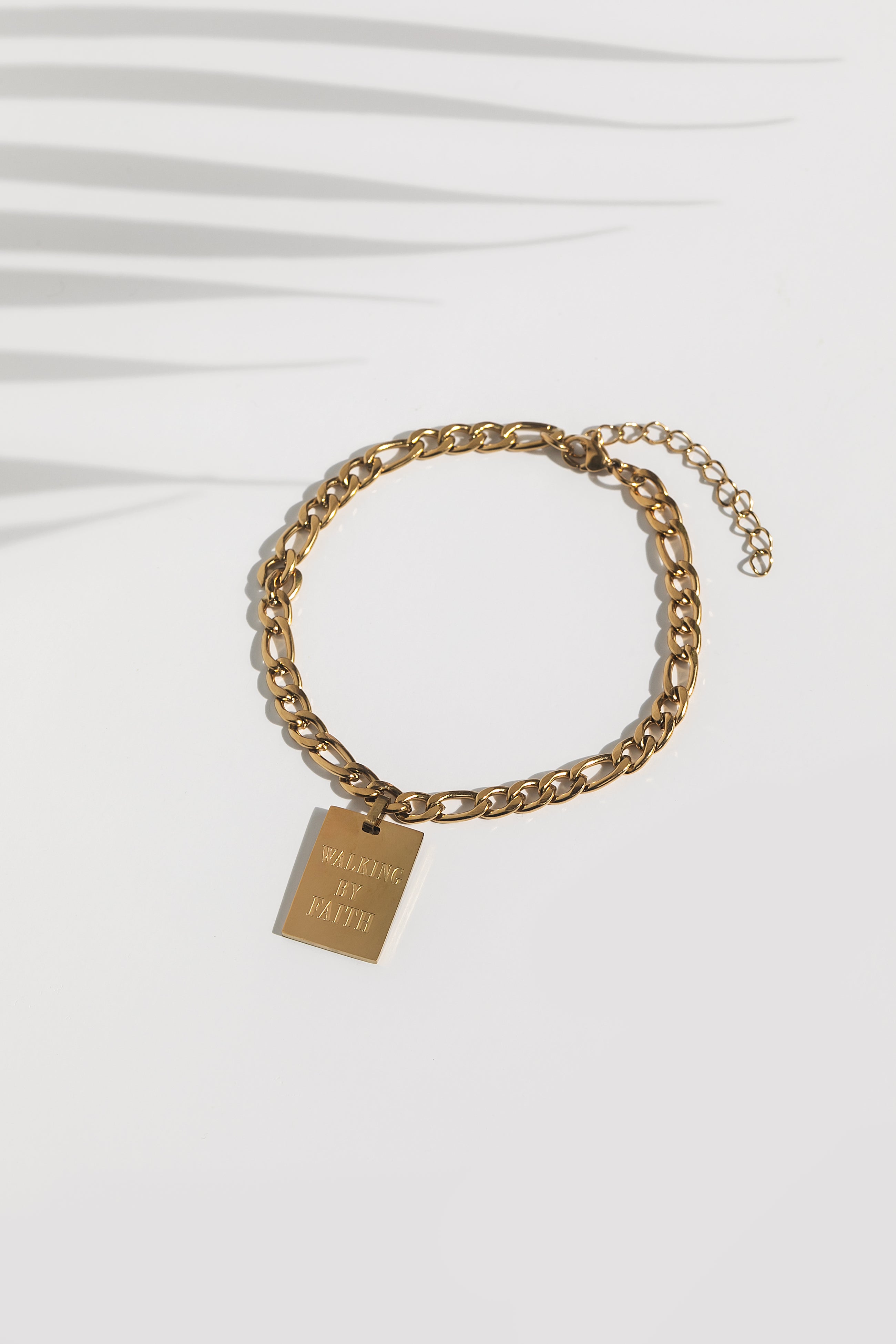 An 18k gold-plated stainless steel anklet with a bold and eye-catching design, featuring a chain link and a rectangular pendant that reads 'WALKING BY FAITH.'.