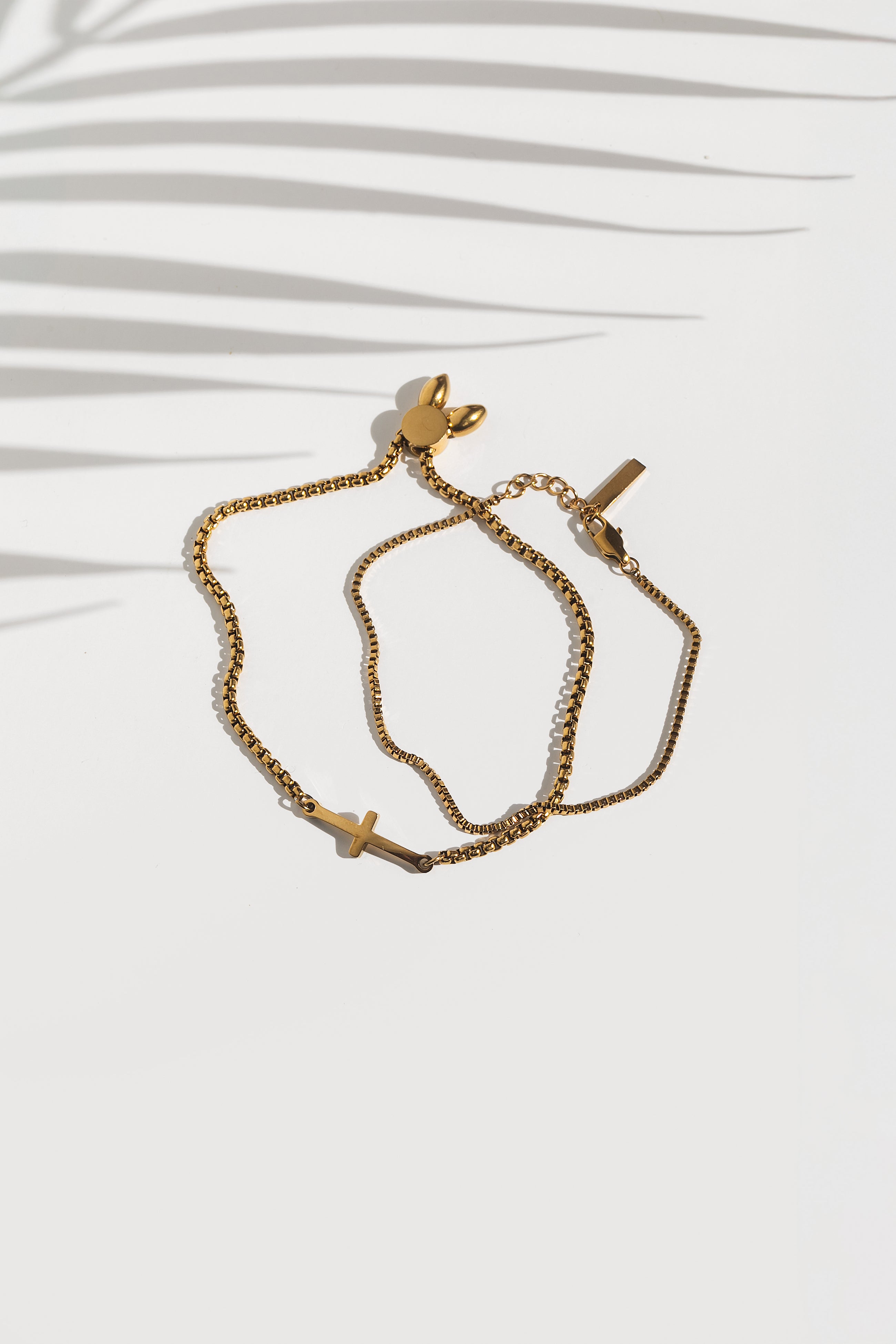 Two gold-plated bracelets, one with a cross charm and one with a plain chain, laid out on a white surface with a shadow cast over them.