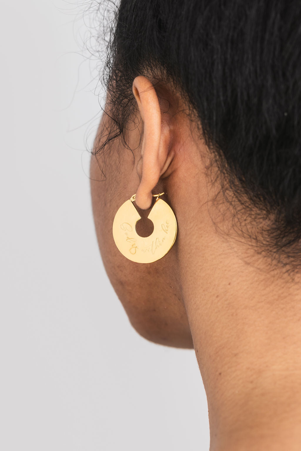 A person wearing 18k gold-plated hoop earrings with an engraved design "God is within her".