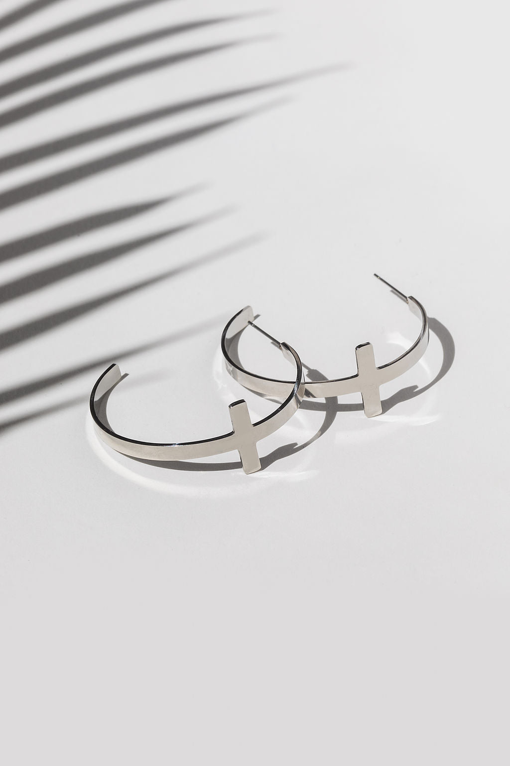 A pair of stainless steel cross hoop earrings on a white surface, with a shadow of a plant leaf cast across them.
