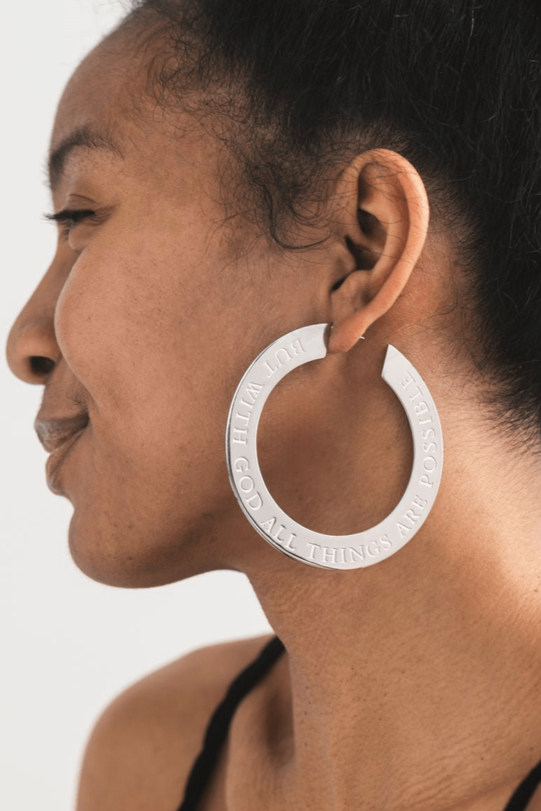 Silver hoop earring engraved with "But with God all things are possible" worn by a woman on a white background