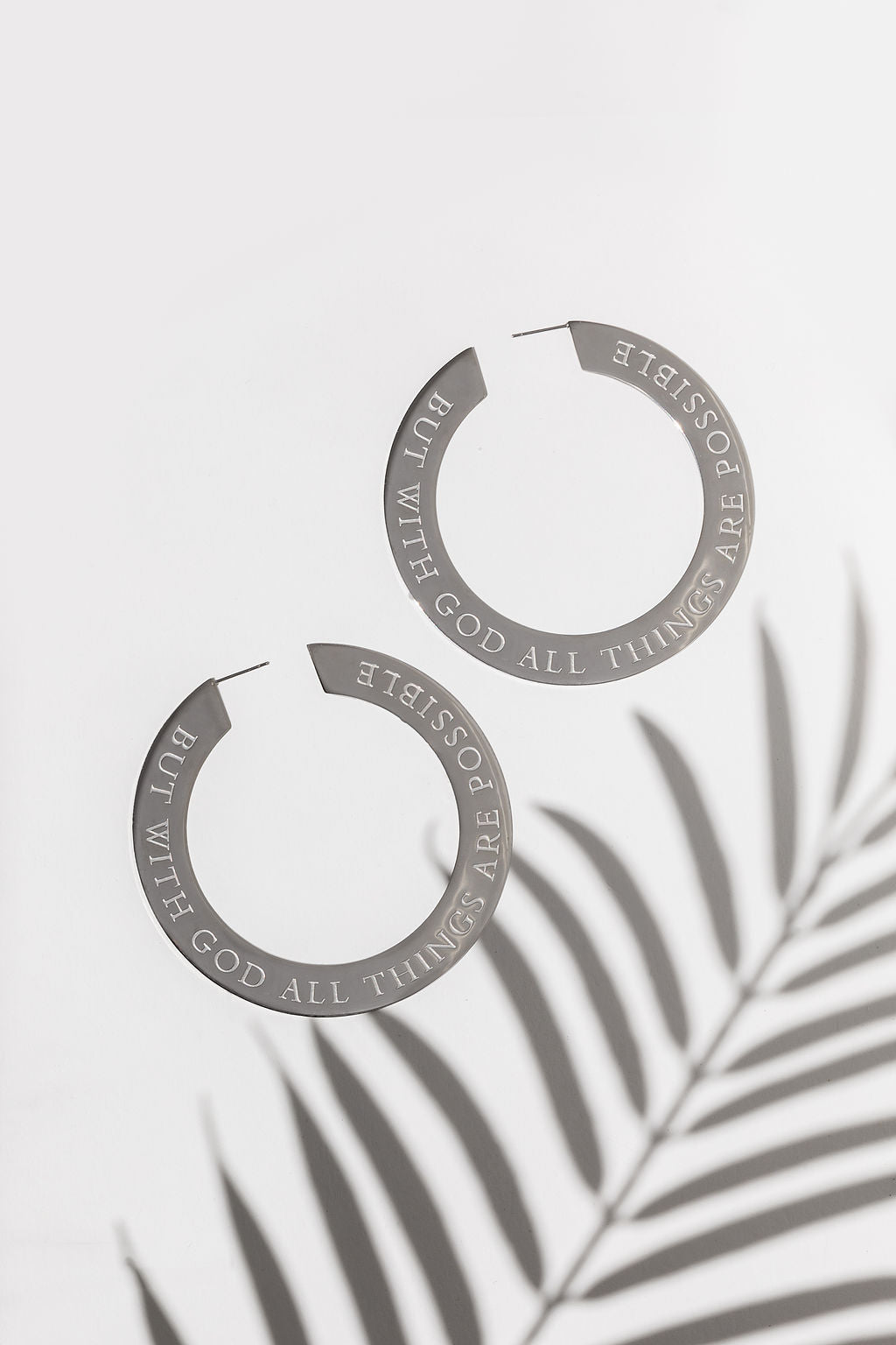 A pair of large stainless steel hoop earrings engraved with "But with God all things are possible" displayed on a white background with a shadow of a plant leaf. The earrings have text engraved on them.