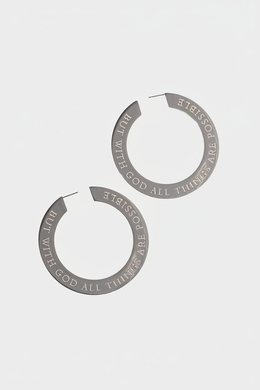 A pair of large stainless steel hoop earrings engraved with "But with God all things are possible" displayed on a white background with a shadow of a plant leaf. The earrings have text engraved on them.