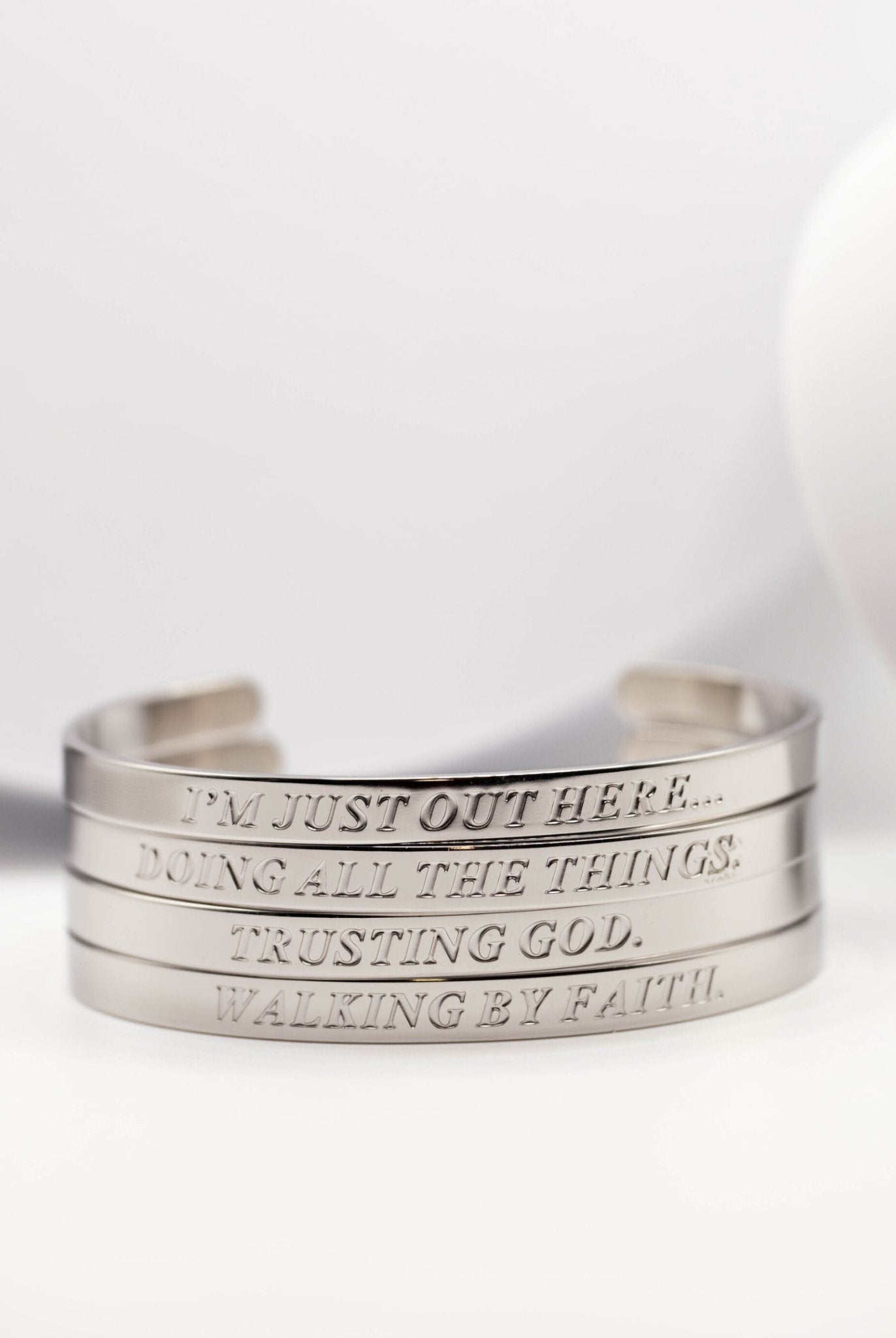 Close-up of four engraved stainless steel Christian bangles stacked together with phrases like “Trusting God” and “Walking by Faith” – inspirational Christian jewelry for women.