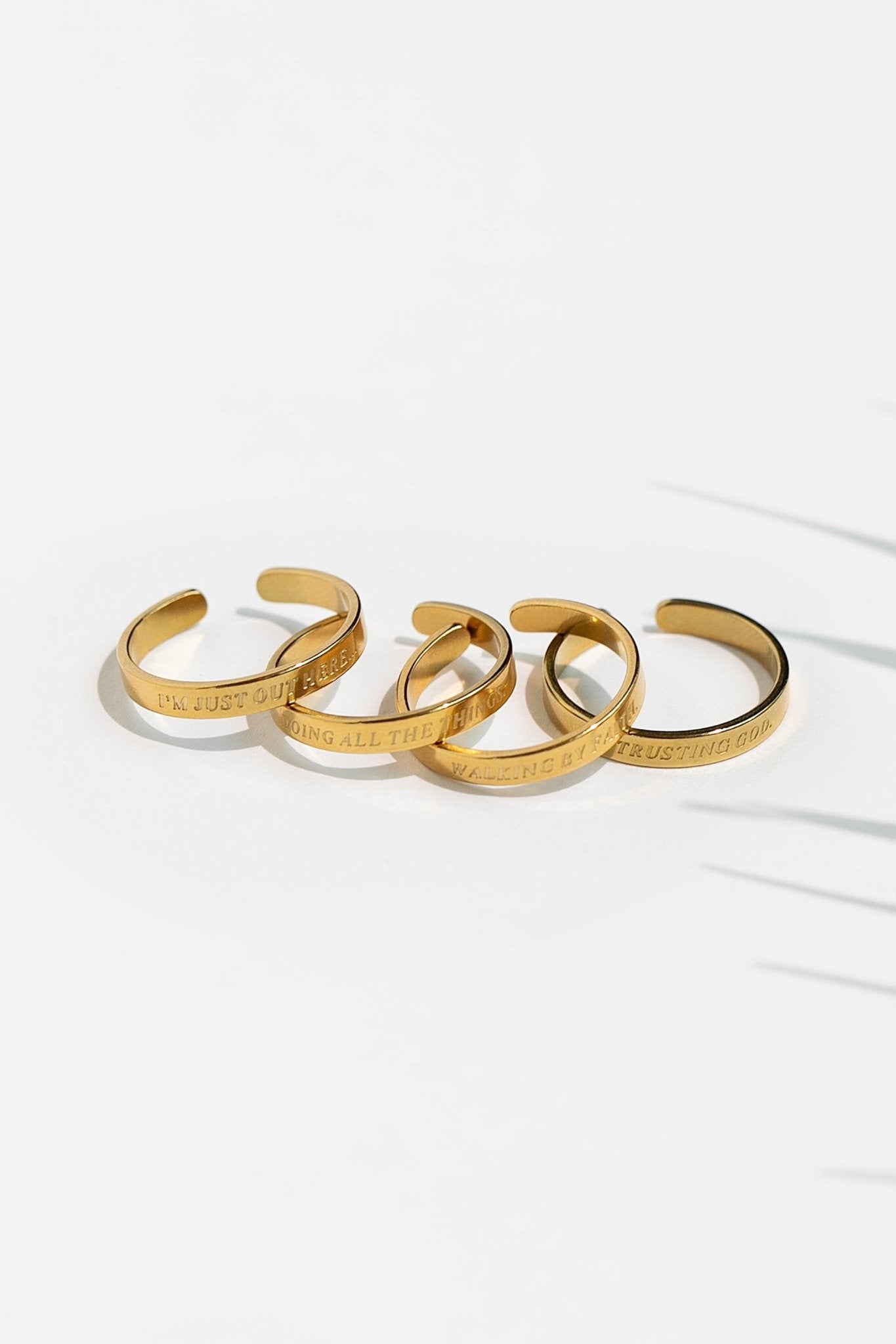 Set of four open-style 18K gold-plated rings engraved with phrases "I'm just out here...", “Walking by Faith”, "Trusting God" and “Doing All the Things,” photographed in bright, minimal light for A Meaningful Mood.
