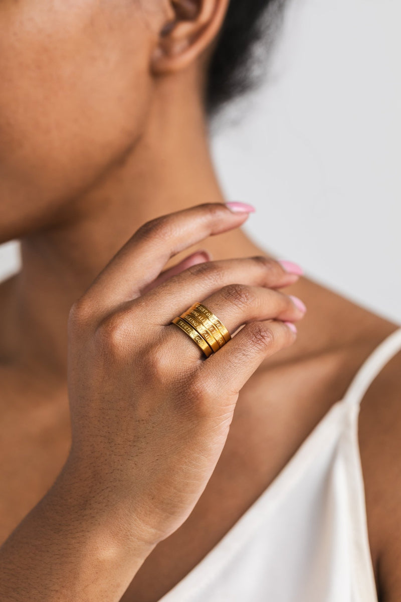 Close-up of a woman’s hand with manicured nails wearing all four gold engraved rings stacked together from A Meaningful Mood’s Faith Reflections Ring Set.