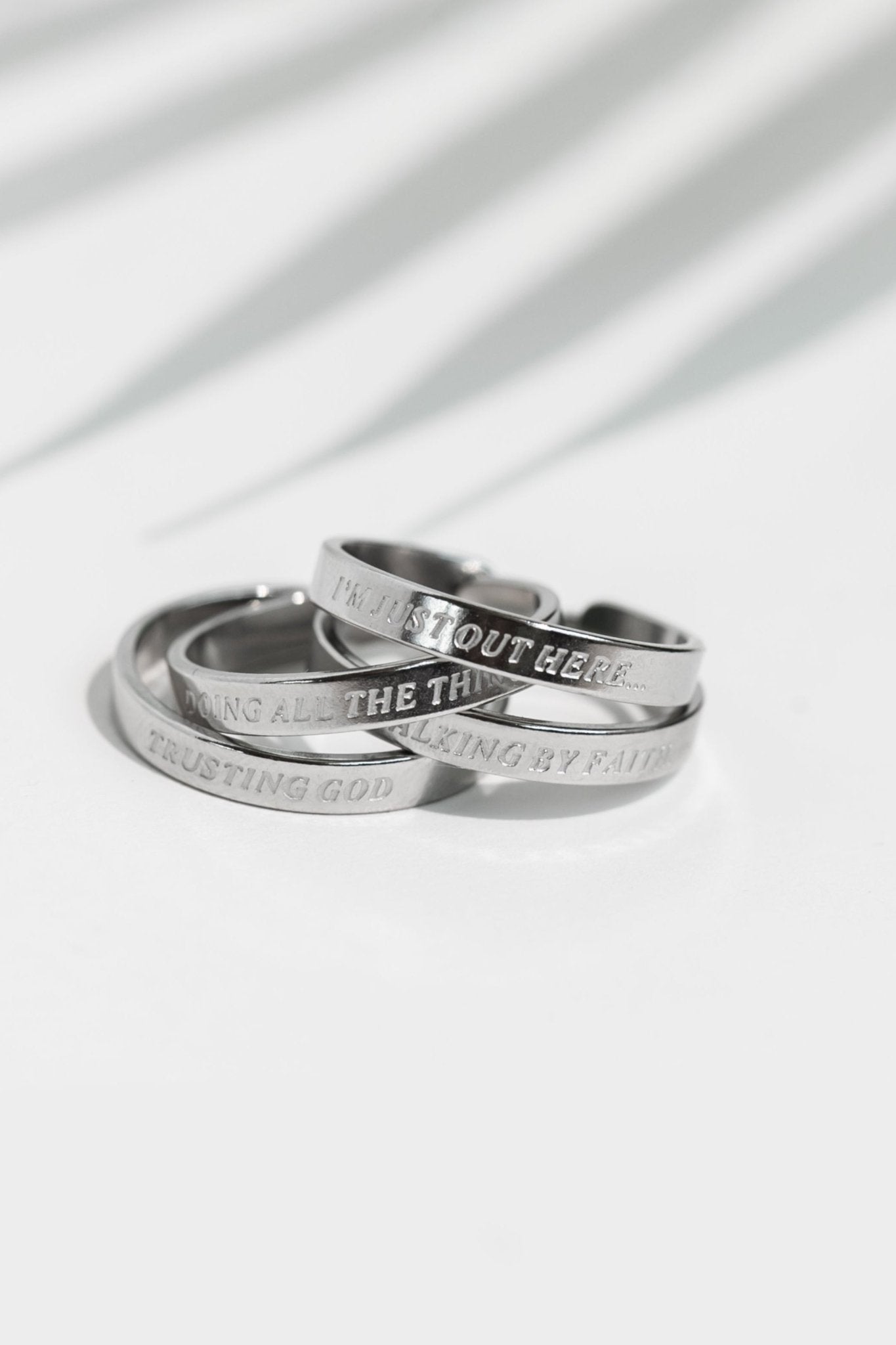 Stack of silver stainless steel rings engraved with “I’m Just Out Here…,” “Doing All the Things,” “Walking by Faith,” and “Trusting God,” part of the Faith Reflections Ring Set from A Meaningful Mood.
