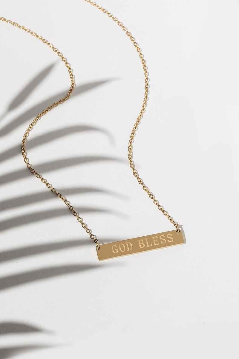 Flatlay of a gold Christian necklace with “God Bless” engraved on a delicate bar pendant – faith-inspired minimalist jewelry for women.