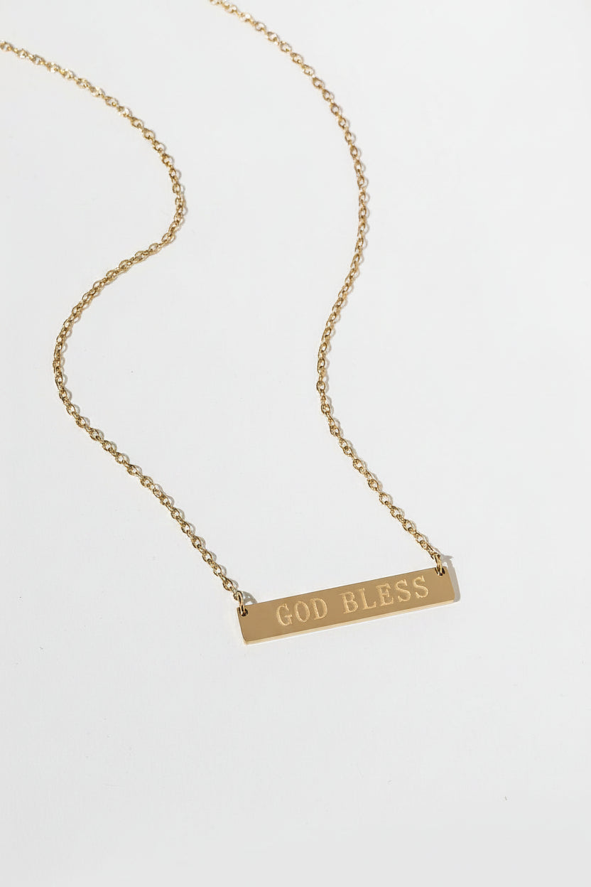 Flatlay of a gold Christian necklace with “God Bless” engraved on a delicate bar pendant – faith-inspired minimalist jewelry for women.