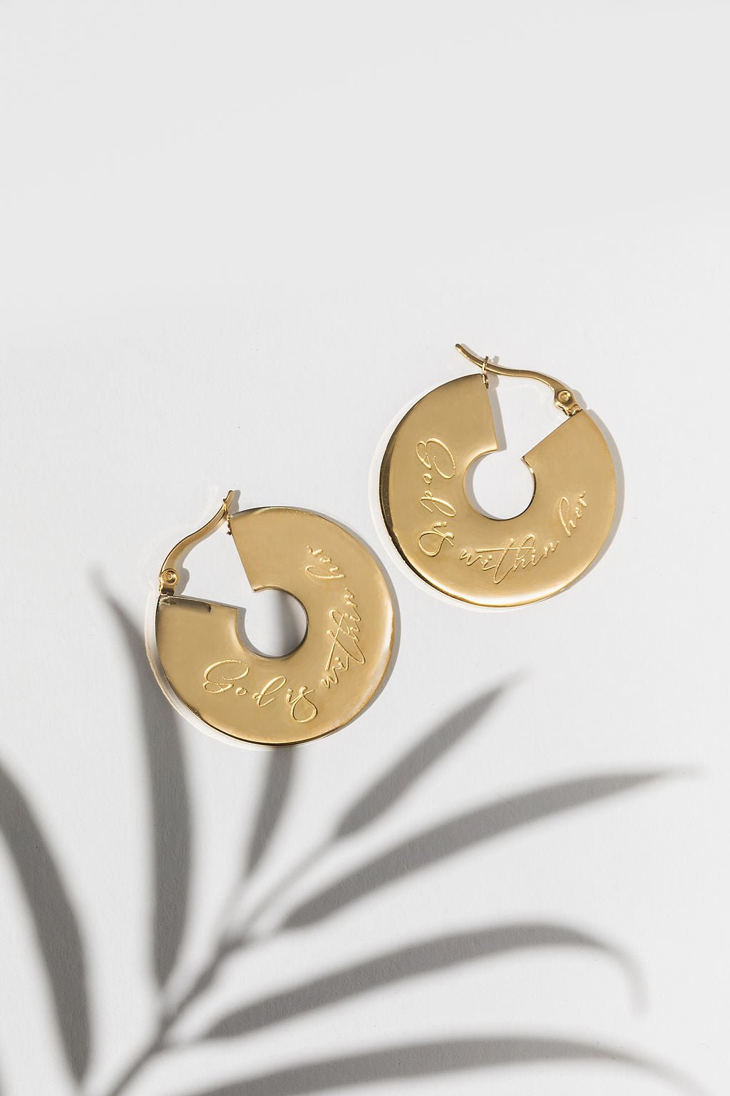 A pair of small 18k gold-plated hoop earrings with an engraved design "God is within her", displayed against a white background with a shadow of a plant to the side.