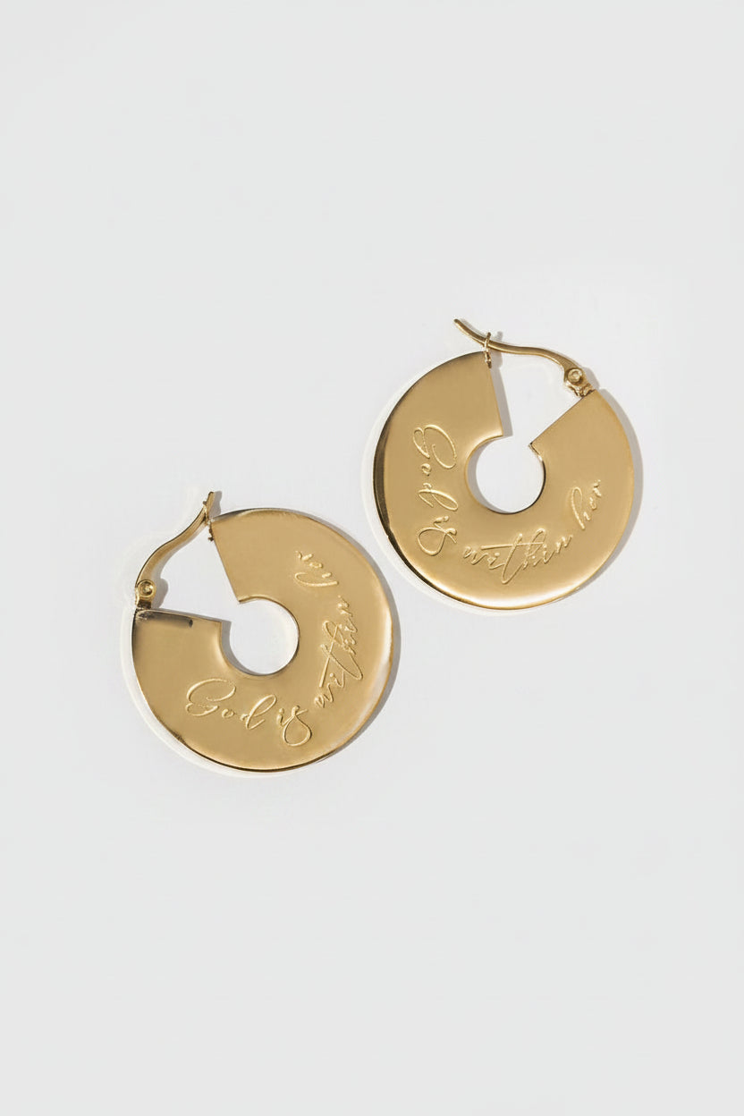 A pair of small 18k gold-plated hoop earrings with an engraved design "God is within her", displayed against a white background with a shadow of a plant to the side.