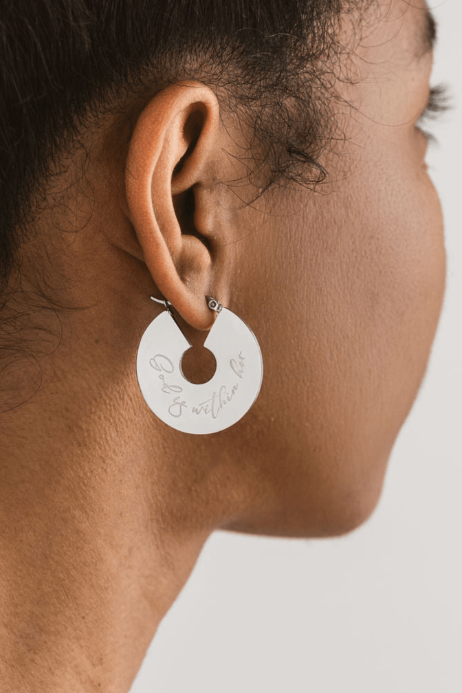 Side profile close-up of a woman wearing a silver earring engraved with "God is within her" on a neutral background.