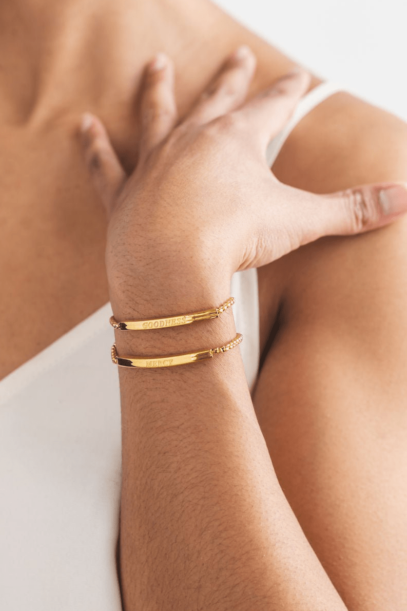 Close-up of a woman’s wrist wearing the Goodness & Mercy 2pc 18k gold plated Christian bracelet set from A Meaningful Mood, engraved with "GOODNESS" and "MERCY" — elegant faith-based jewelry with scripture-inspired meaning.