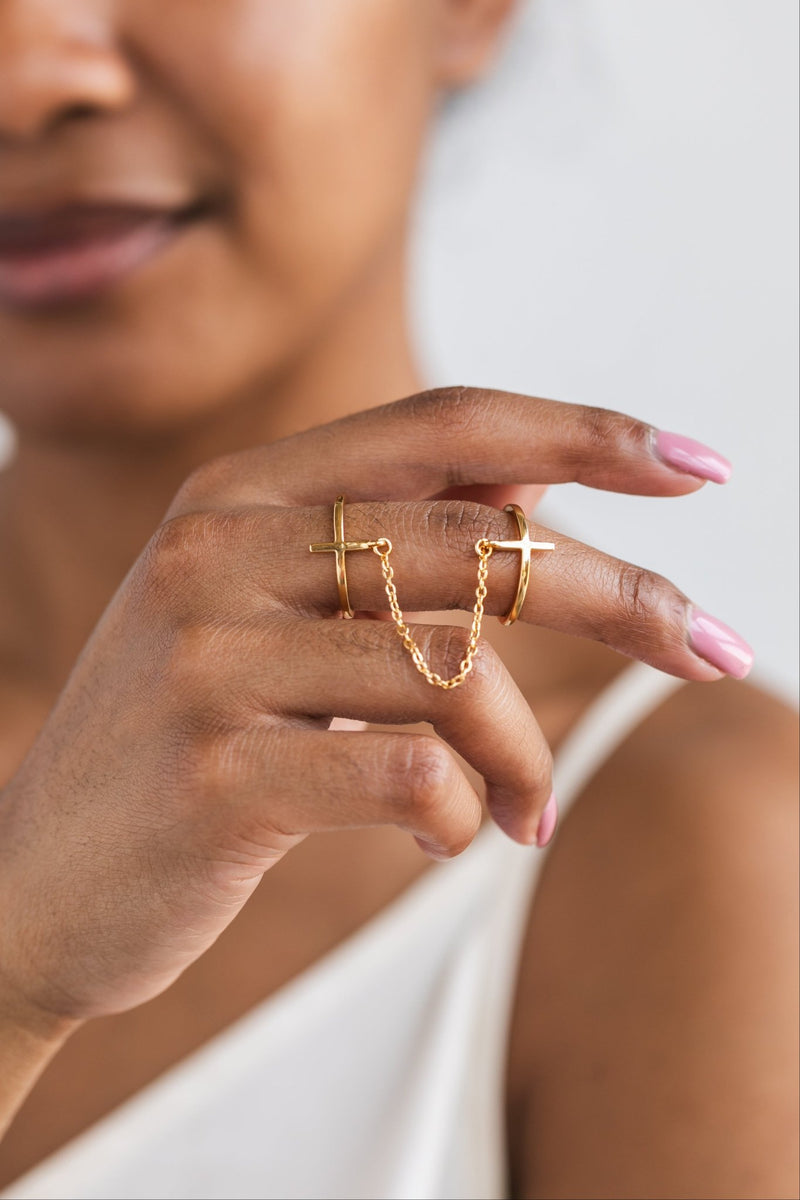 Front-facing close-up of a woman’s hand wearing the So Chic Chain Cross Rings across stacked on one finger, highlighting the delicate gold chain and minimalist cross detail.