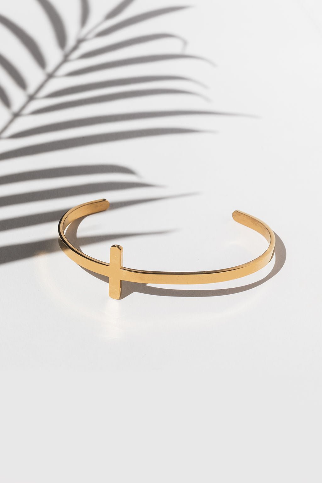 Minimalist 18k gold plated bangle featuring a bold cross detail, photographed against a white backdrop with leaf shadows.