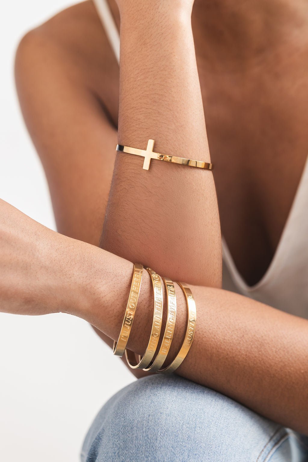 Close-up of a woman wearing a stack of four gold bangles engraved with scripture phrases, paired with a sleek gold cross bangle on the other wrist.