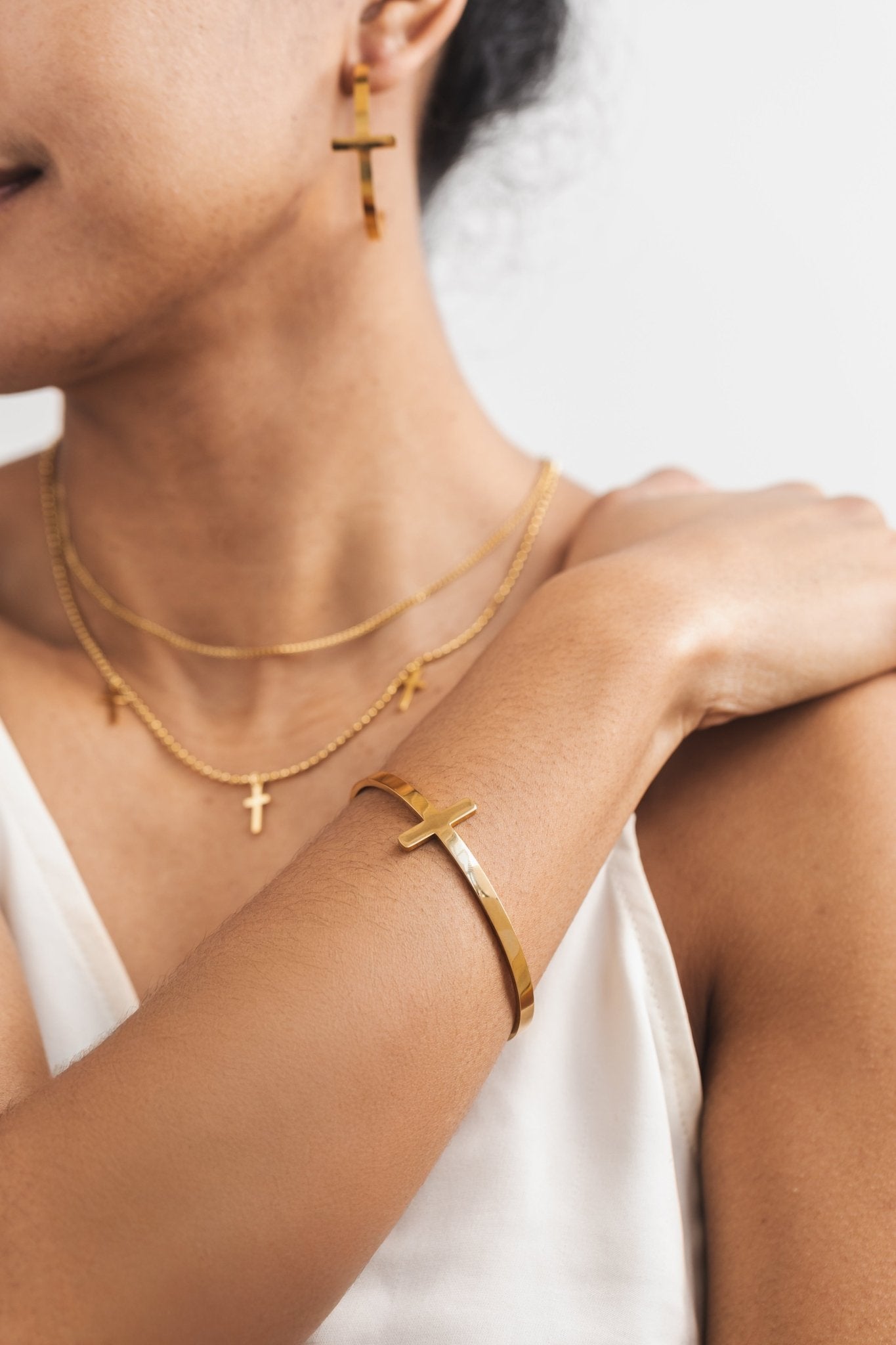 Woman wearing a gold cross bangle, matching layered cross necklace and matching open hoop cross earrings.