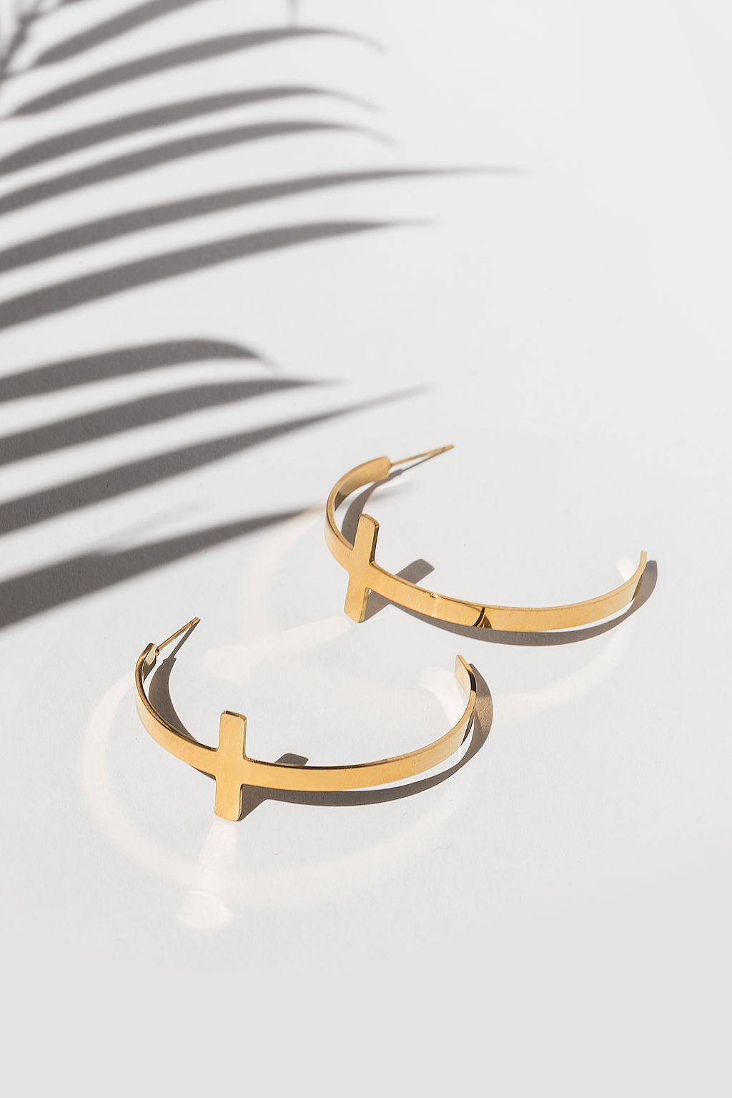 Flat lay of A Meaningful Mood’s So Chic Cross Hoops in 18k gold plated stainless steel with soft palm shadows. Lightweight, faith-inspired Christian cross earrings that blend faith and fashion.