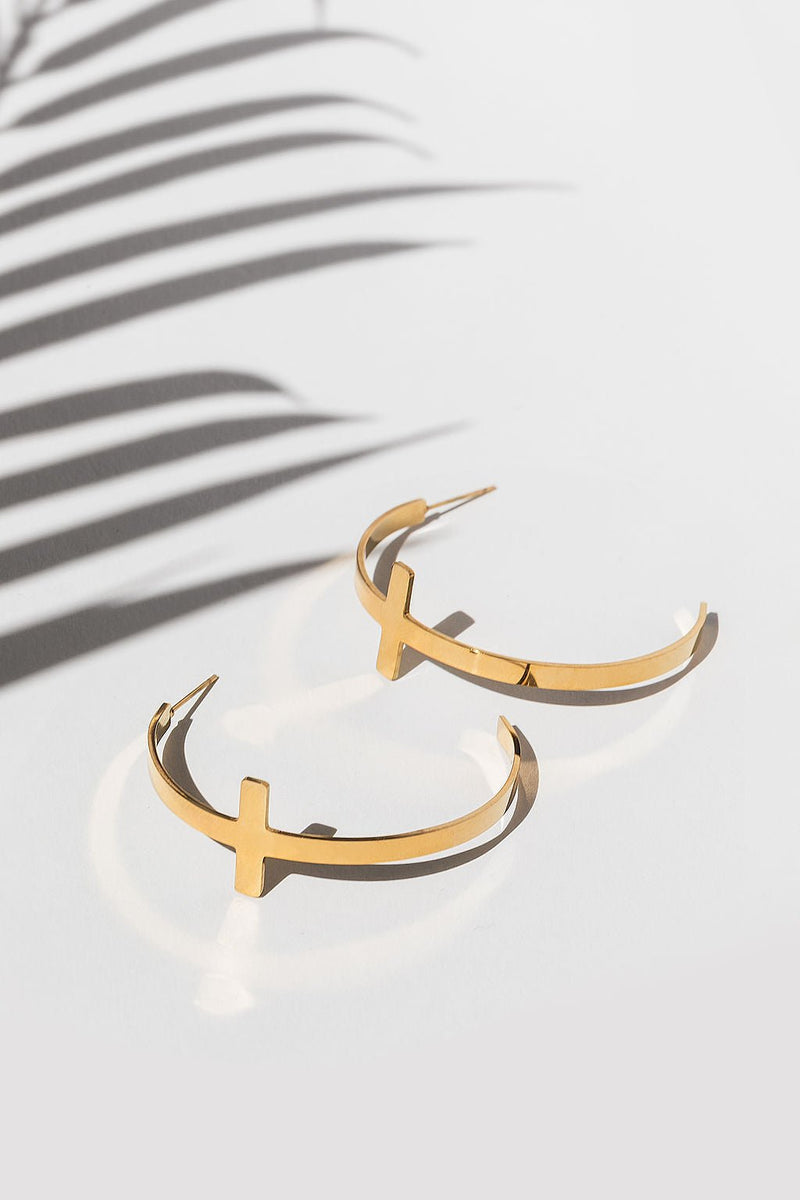 Flat lay of A Meaningful Mood’s So Chic Cross Hoops in 18k gold plated stainless steel with soft palm shadows. Lightweight, faith-inspired Christian cross earrings that blend faith and fashion.