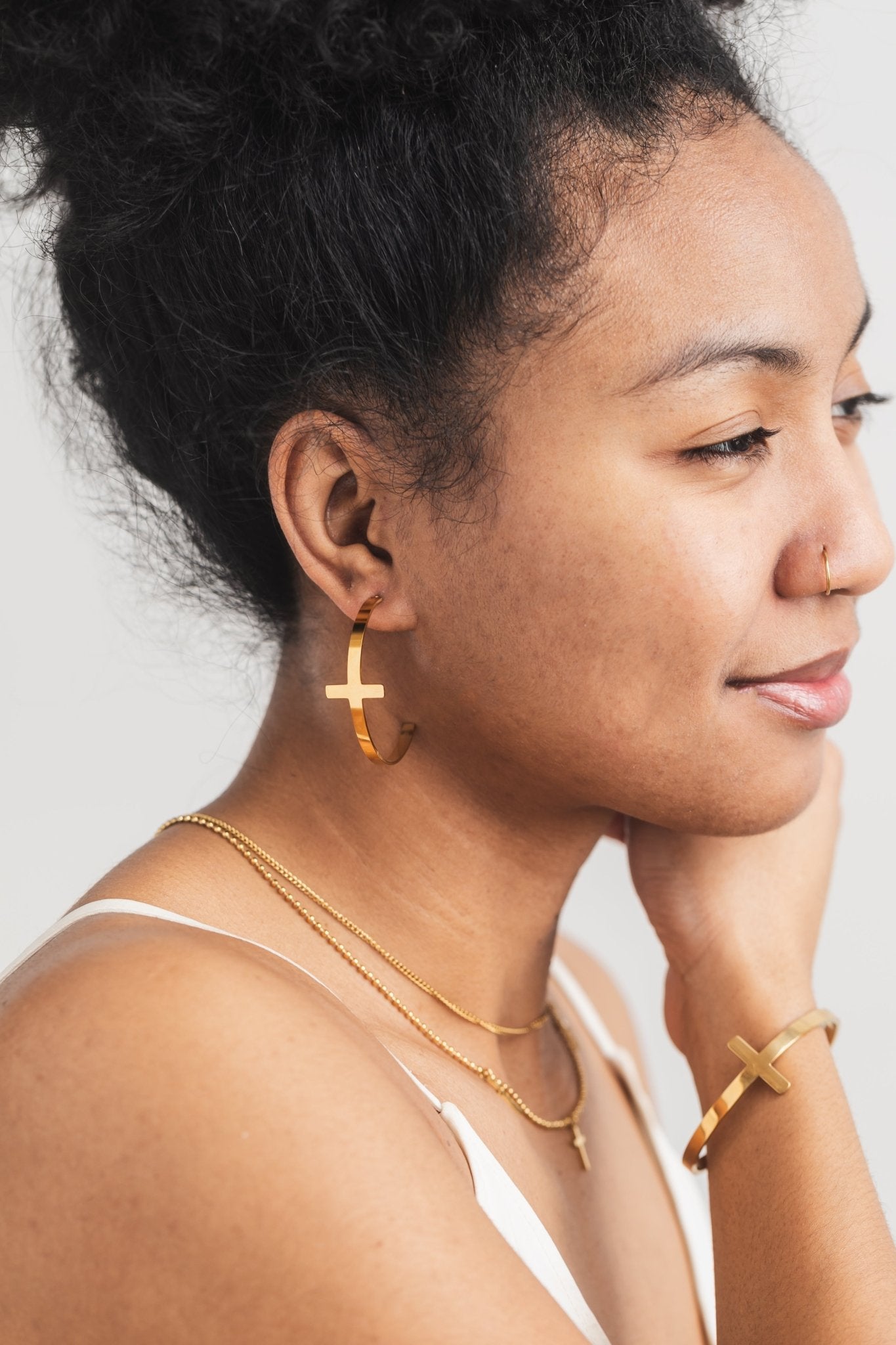 Close-up side view of a woman wearing A Meaningful Mood's So Chic Cross Hoops in 18k gold plated stainless steel, paired with matching layered gold cross necklaces and bangle bracelet. Christian statement jewelry for stylish women who reflect their faith through fashion.