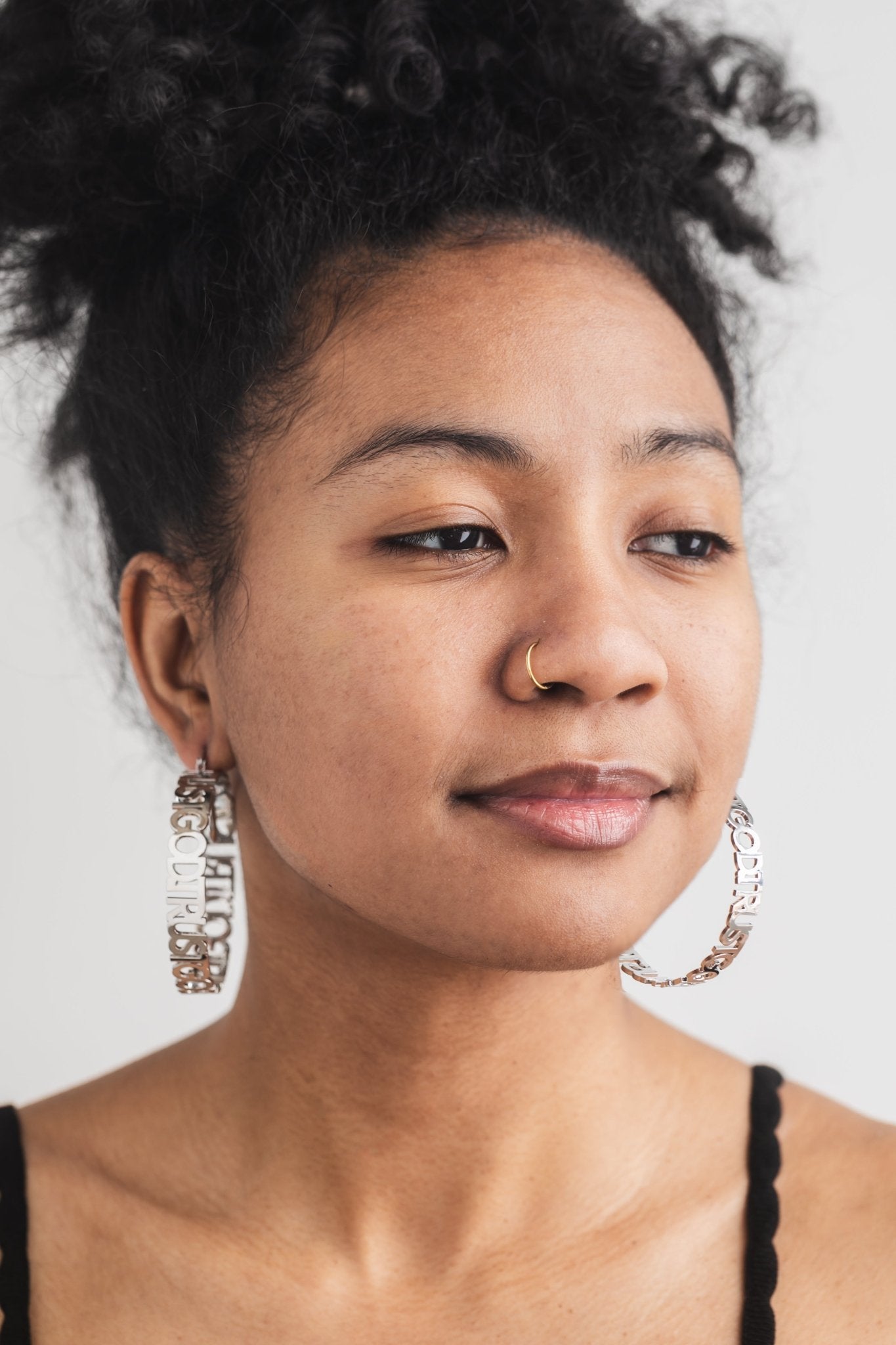 Close-up of woman wearing silver stainless steel "Trust God" hoop earrings from A Meaningful Mood. Christian statement earrings styled with natural makeup and gold nose ring. Faith-based jewelry for women who reflect their faith and style.