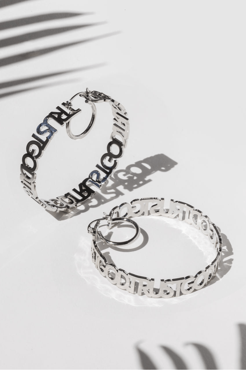 Flatlay of silver stainless steel "Trust God" hoop earrings on white background with shadow detail. Faith-inspired Christian jewelry from A Meaningful Mood, perfect for stylish women of faith seeking meaningful accessories.