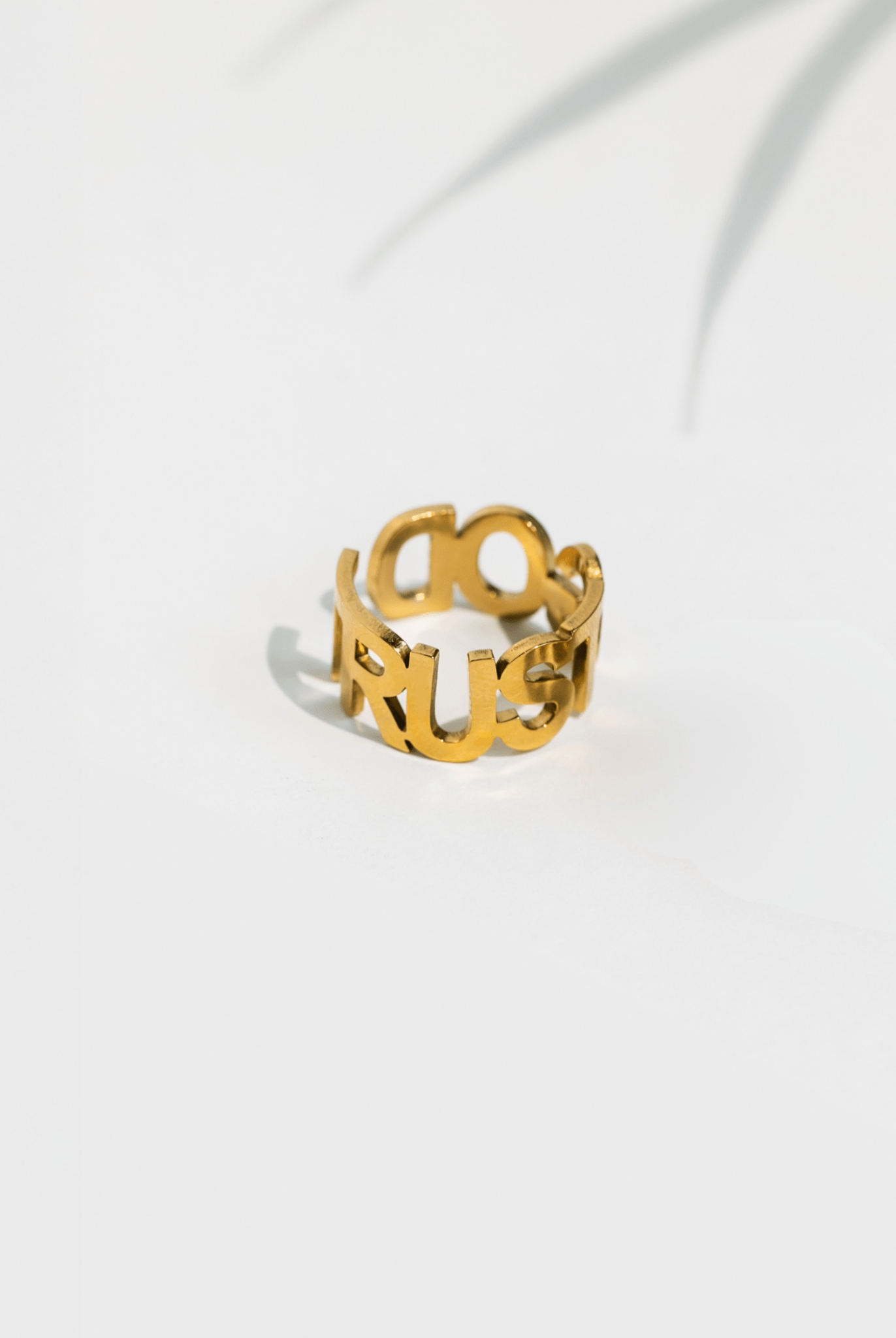 Gold-plated adjustable ring with cutout “TRUST GOD” lettering, photographed upright with soft shadow and palm leaf detail in background.