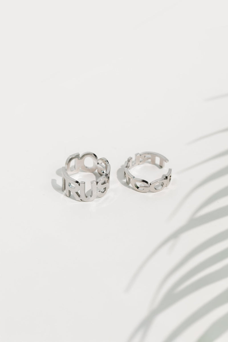 Two stainless steel Trust God rings side by side on a white surface, showing both the “TRUST” and “GOD” sides of the bold cutout design in polished silver tone.