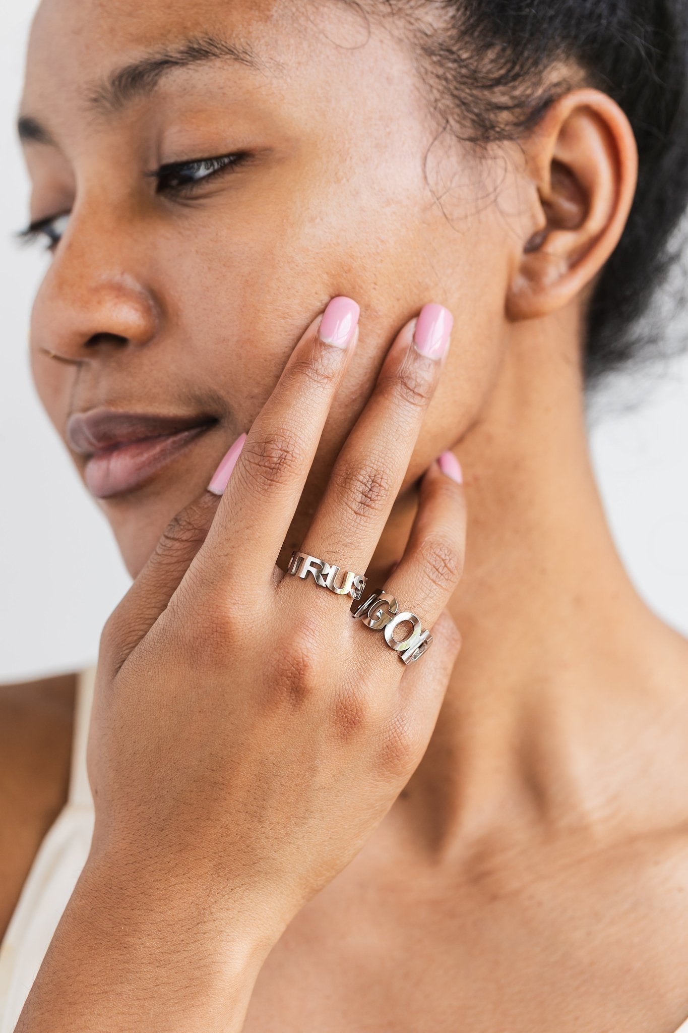 Close-up of a woman with pink nails gently touching her face, wearing the TRUST GOD stainless steel rings on two fingers highlighting the two sizing styles.