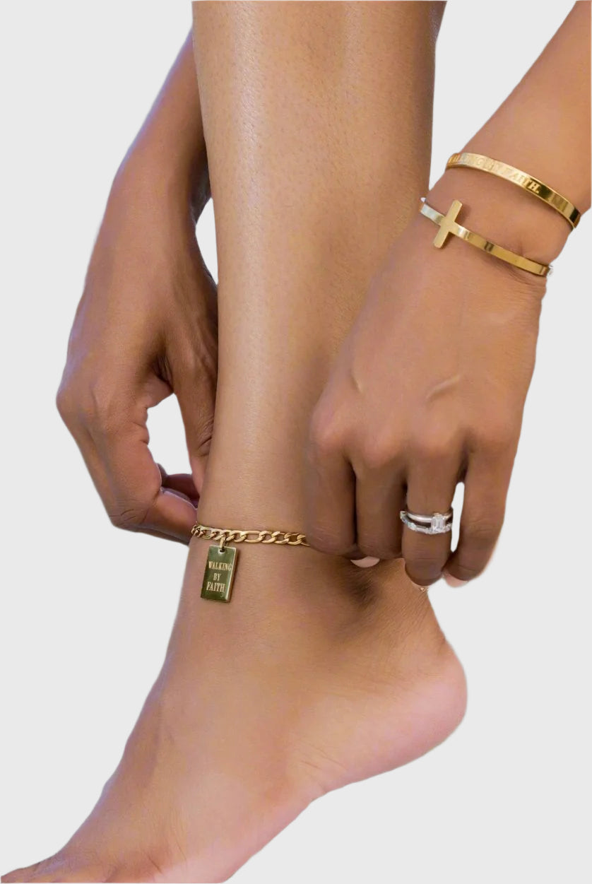 Solo shot of a gold chain anklet with a rectangular "Walking by Faith" engraved pendant, laid flat on a white surface with soft palm leaf shadows.
