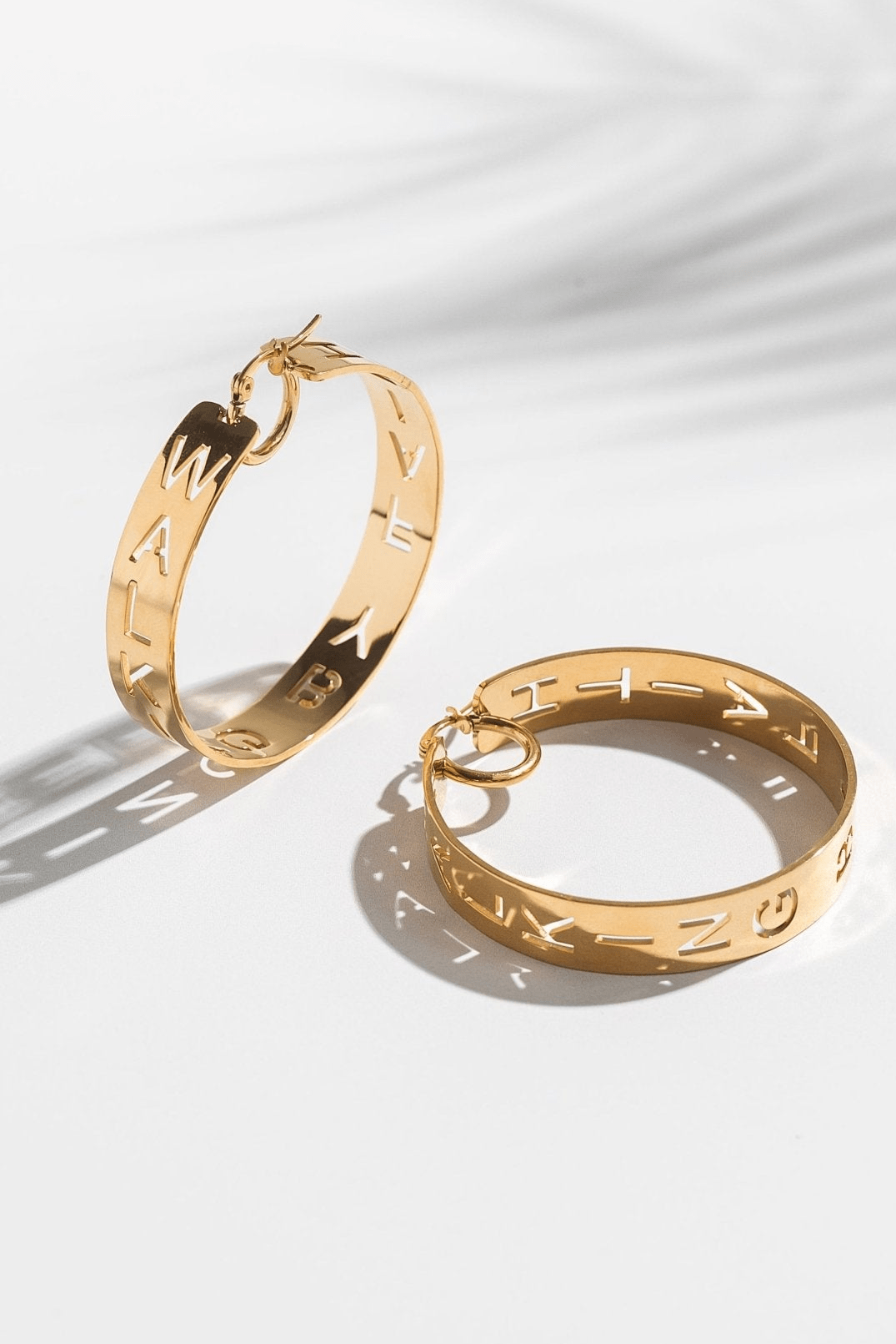 Pair of gold statement hoop earrings with cutout design featuring "Walking by Faith" displayed on a white surface. Christian jewelry by A Meaningful Mood.