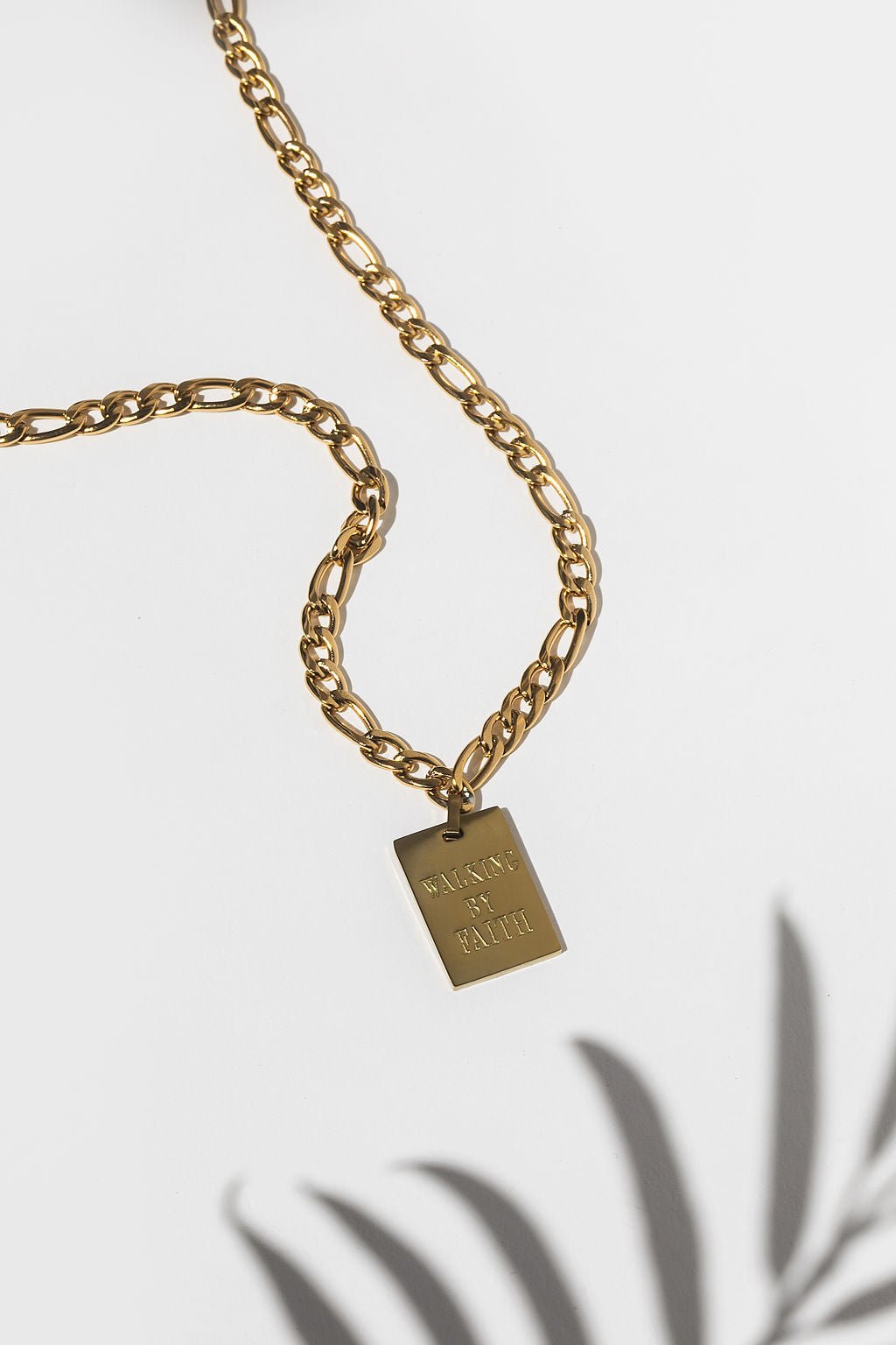 Flat lay of the Walking By Faith Pendant Necklace featuring an 18k gold plated figaro chain with a rectangular charm engraved with 'WALKING BY FAITH,' displayed on a white background with soft palm shadows.