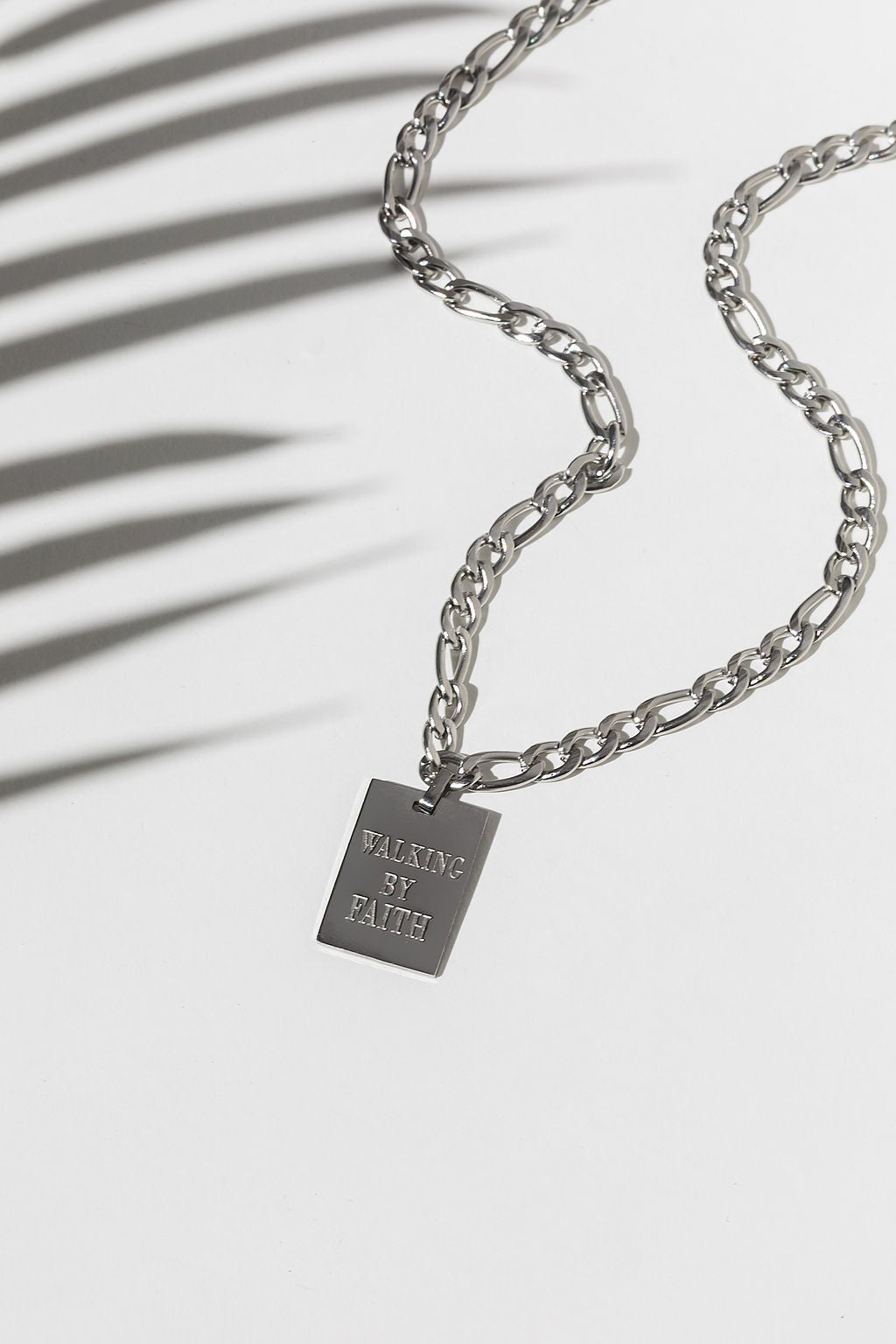 A stainless steel pendant necklace with a square tag engraved with 'WALKING BY FAITH' laid out on a white surface with a shadow of a plant leaf.