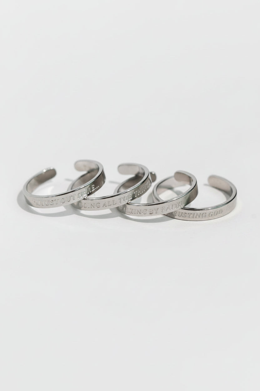 Four silver stainless steel engraved rings laid flat in soft lighting, each inscribed with faith-inspired phrases from the Faith Reflections Ring Set by A Meaningful Mood.