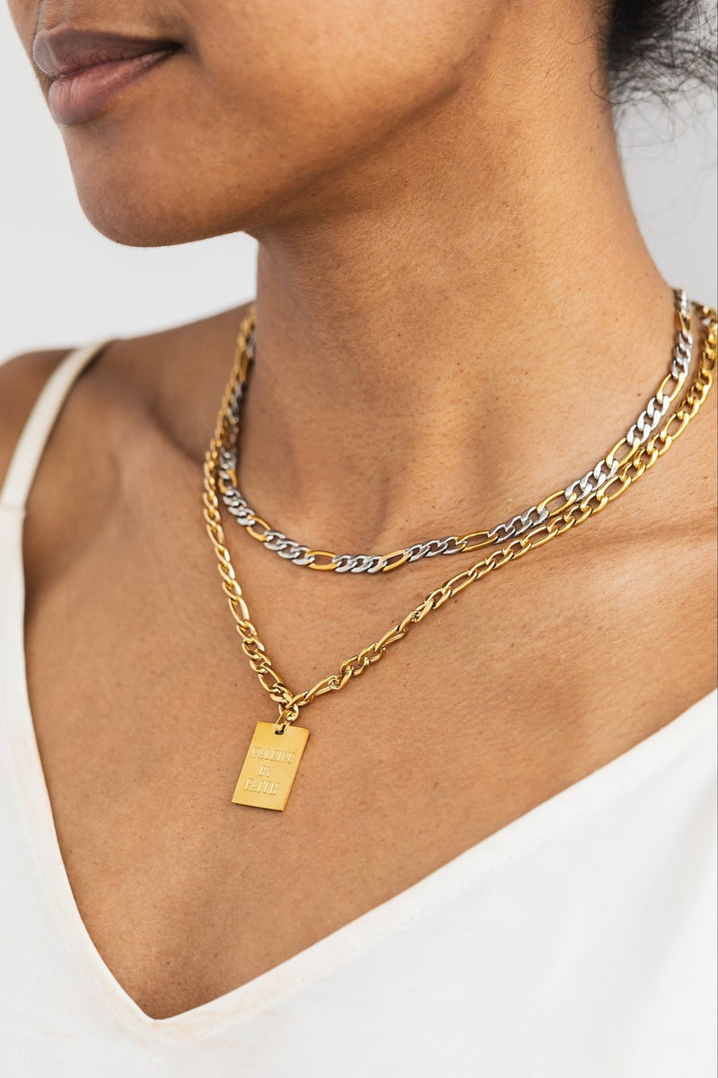 Layered necklace stack featuring the Duo Luxe Two-Tone chain paired with a gold “Walking by Faith” pendant necklace, styled on a woman wearing a white top.