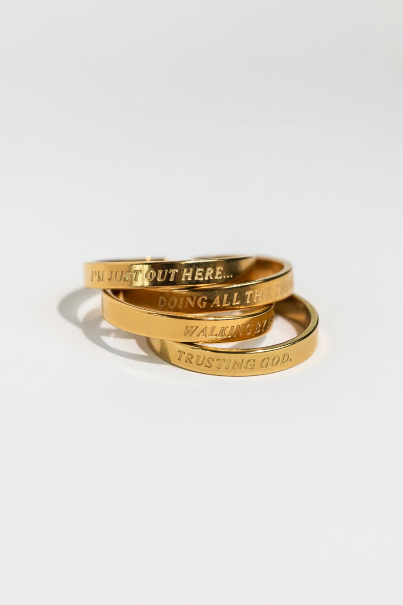 Stack of four 18K gold-plated engraved rings featuring faith-based phrases “I’m Just Out Here…”, "Doing all the things", "Walking by Faith" and “Trusting God,” from the Faith Reflections Ring Set by A Meaningful Mood.