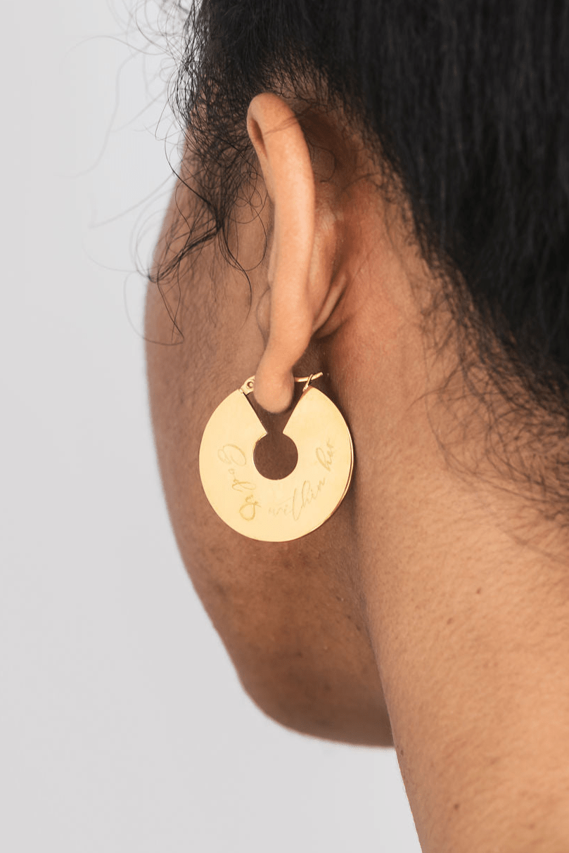 Gold hoop earring engraved with "God is within her" worn by a woman with a plain background