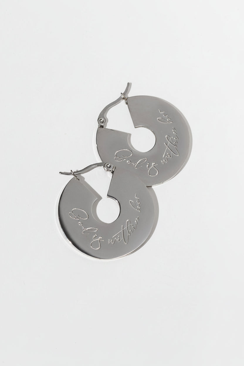 A pair of small engraved hoop earrings inscribed with "God is within her" made of stainless steel, displayed against a light background with a shadow of a plant leaf on them.