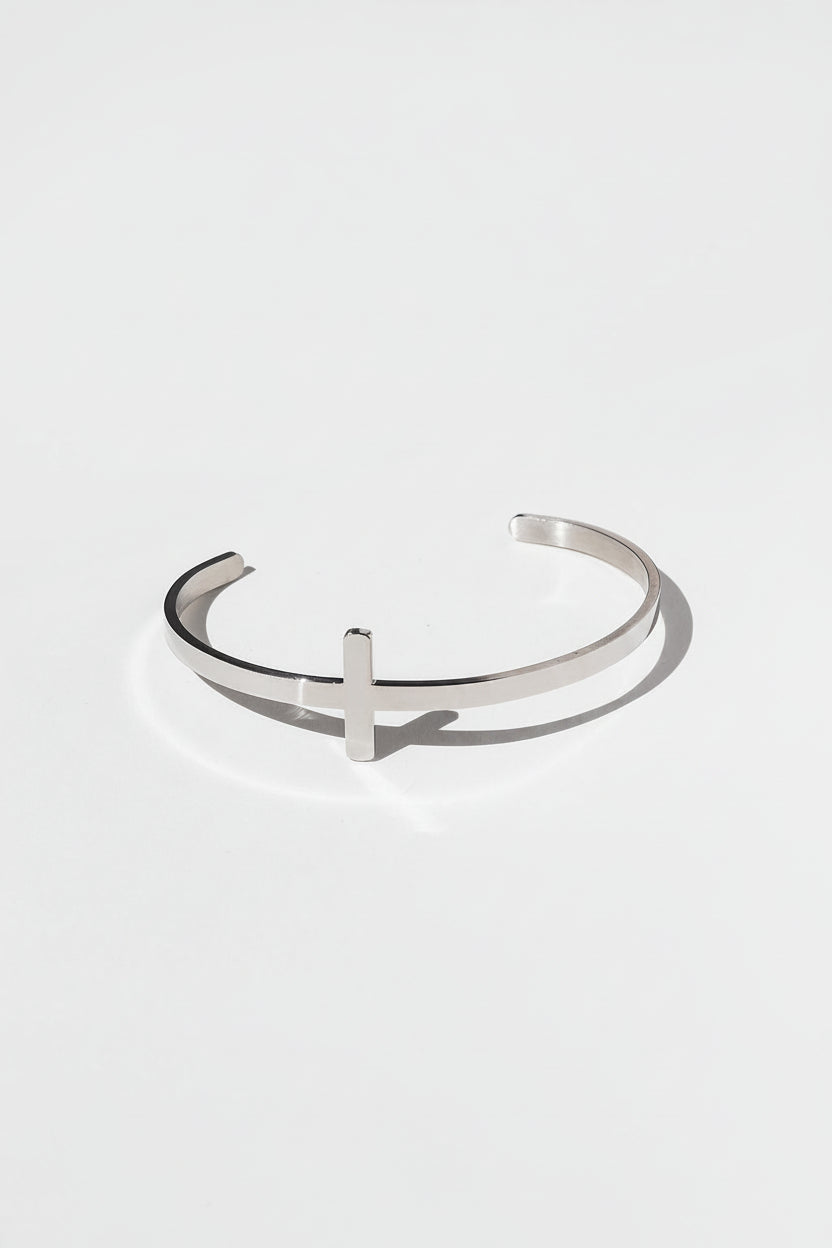 Minimalist stainless steel cross bangle bracelet for Christian women, styled on white background with soft palm shadows.