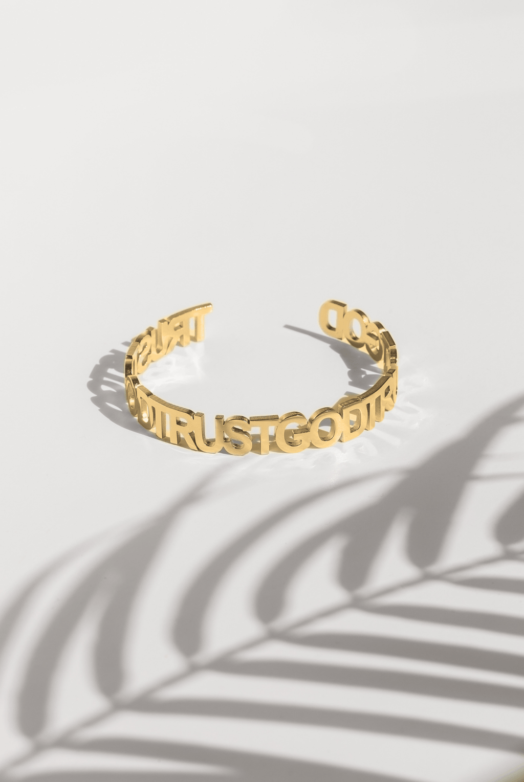 A gold plated stainless steel bangle with the inscription 'TRUST GOD' written around its exterior, displayed against a white background with a shadow of a plant leaf across it.