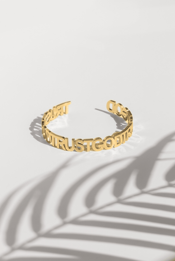 A gold plated stainless steel bangle with the inscription 'TRUST GOD' written around its exterior, displayed against a white background with a shadow of a plant leaf across it.