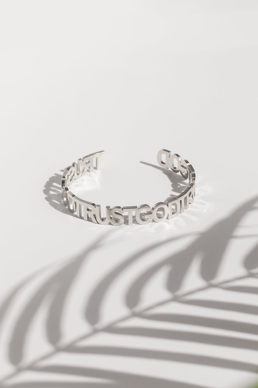 A silver bangle bracelet with the inscription 'Trust God' repeated around it, displayed on a white background with a shadow of a plant leaf over it.