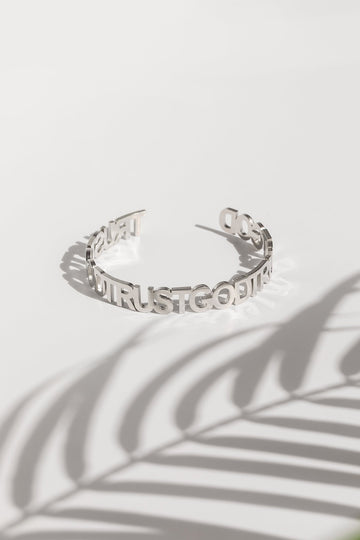 A silver bangle bracelet with the inscription 'Trust God' repeated around it, displayed on a white background with a shadow of a plant leaf over it.