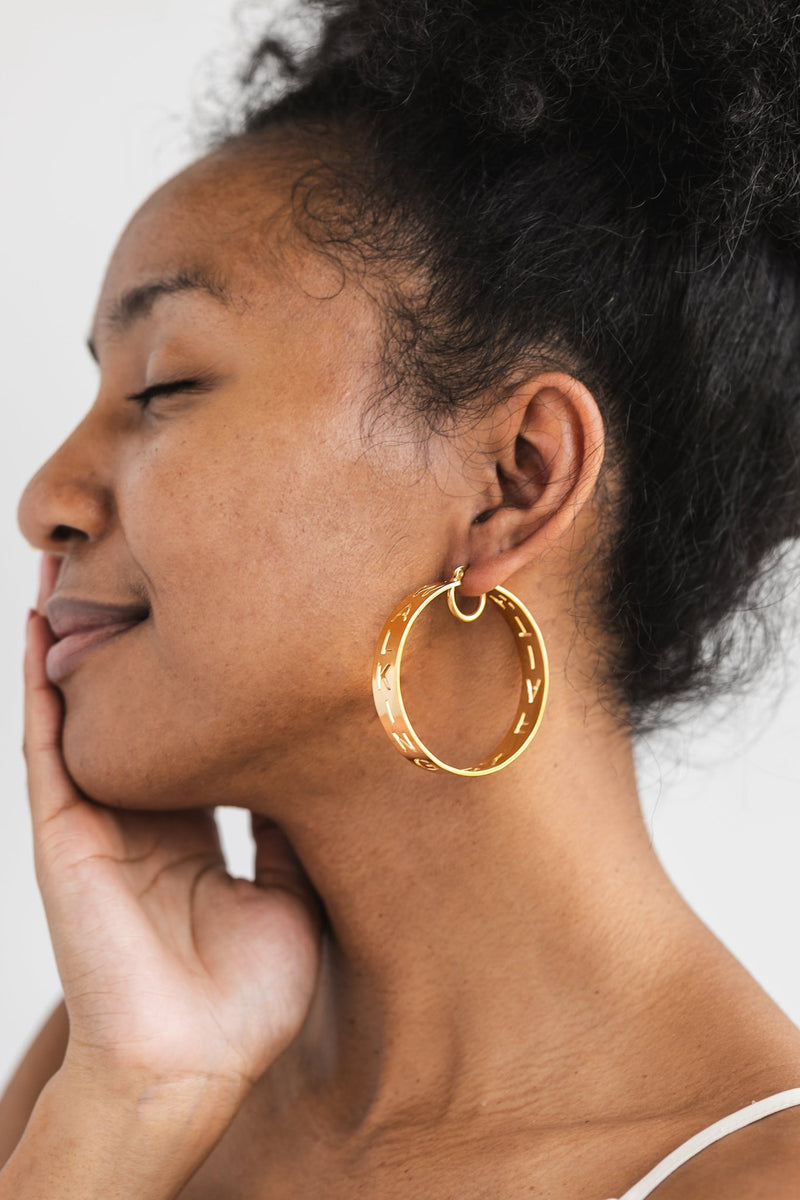 Close-up of a woman smiling with eyes closed, wearing a bold gold hoop earing that says "Walking by Faith" from A Meaningful Mood.
